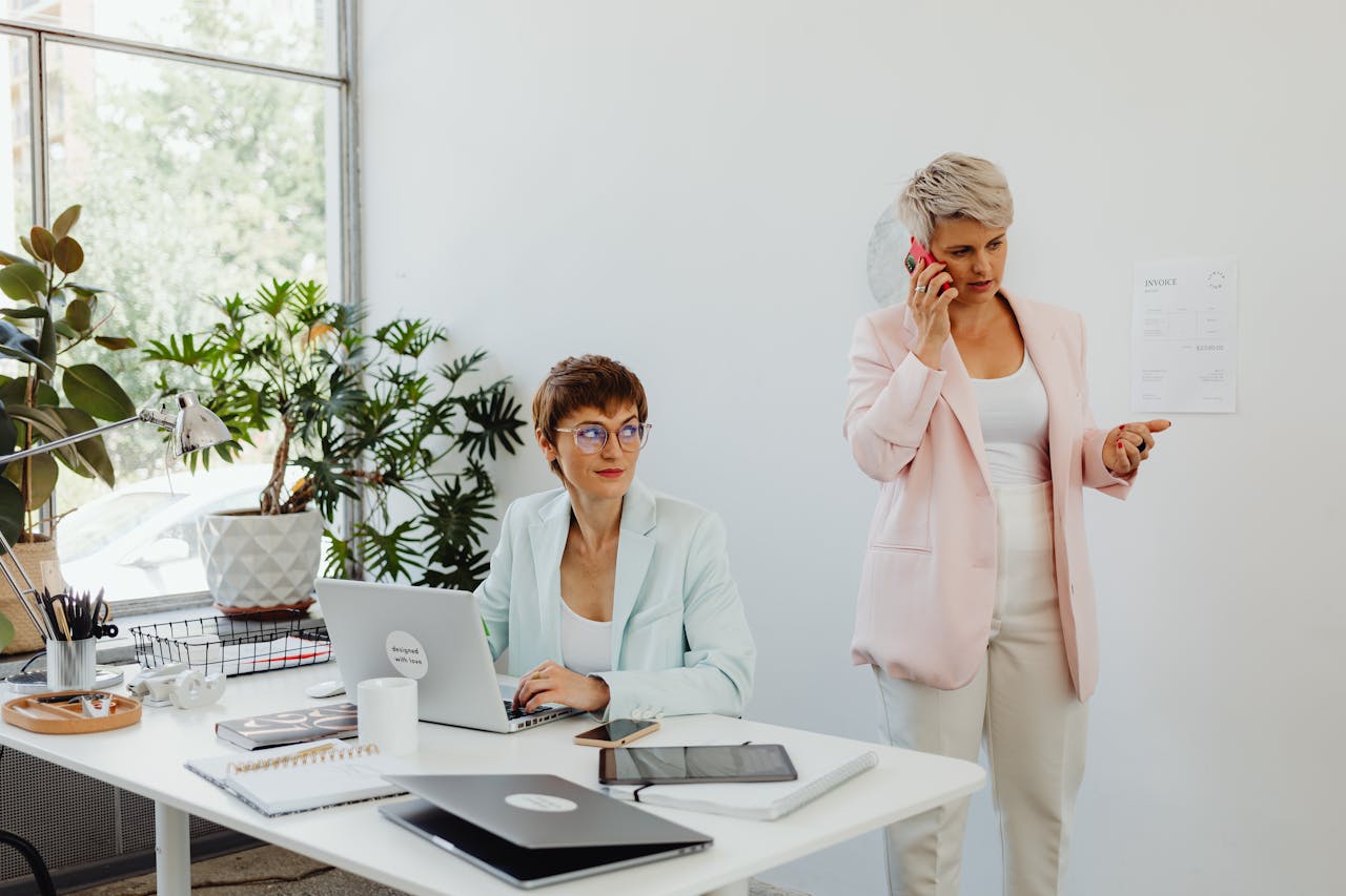 gallery-3 Two businesswomen working together in a modern office setting, using laptops and making phone calls.